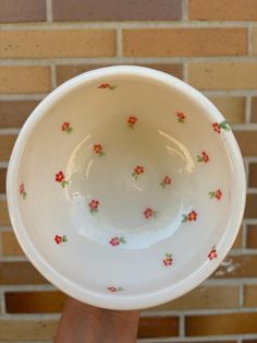 a hand holding a white bowl with red and green flowers on the rim in front of a brick wall