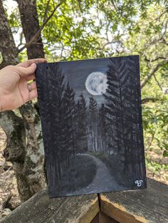 a person holding up a painting with trees in the background and a full moon above it