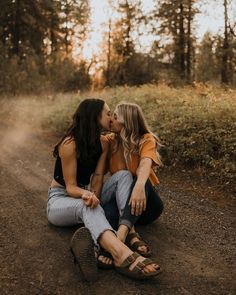 two women sitting on the ground kissing each other in front of trees and dirt road