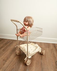 a doll sitting in a shopping cart on top of a hard wood floor next to a white wall