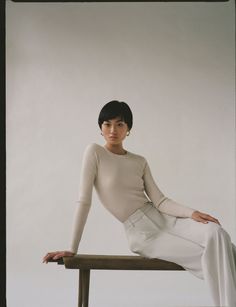 a woman sitting on top of a wooden bench wearing white pants and a turtle neck sweater