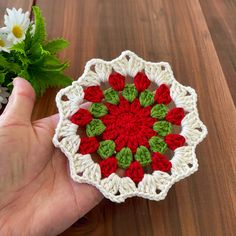 a hand is holding a crocheted doily with red and green flowers