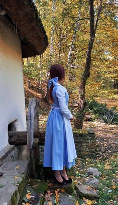 a woman in a blue dress standing next to a white building and looking into the woods