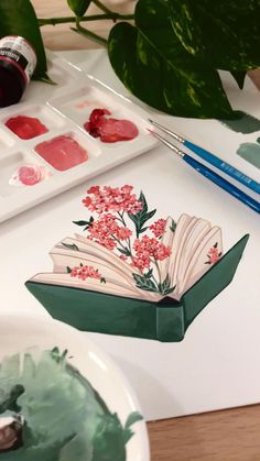 an open book sitting on top of a table next to paintbrushes and watercolors