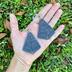 a hand is holding two triangular shaped earrings in front of some green grass and leaves