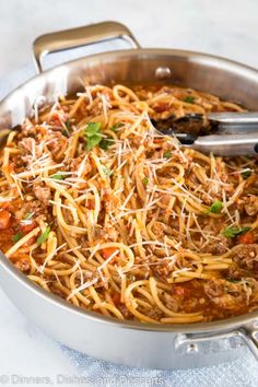 a pan filled with spaghetti and meat on top of a white tablecloth next to a silver spoon