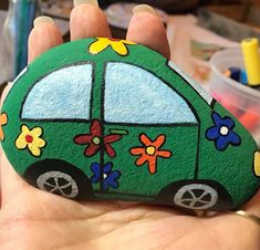 a hand holding a painted rock with a green car on it's side and flowers all over the top