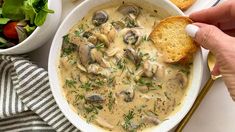 a person dipping some bread into a bowl of mushroom soup with salad in the background