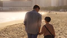 a father and son walking on the beach at sunset with buildings in the back ground