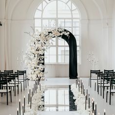 an aisle decorated with white flowers and black chairs in front of a large arched window