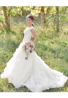 a woman in a wedding dress is standing in the grass and holding a flower bouquet