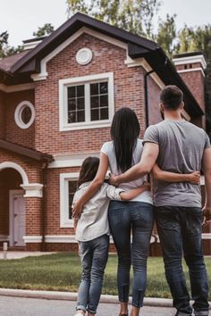 three people standing in front of a house with their arms around each other's shoulders