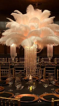 a tall centerpiece with white feathers on top of a black table cloth and gold place settings