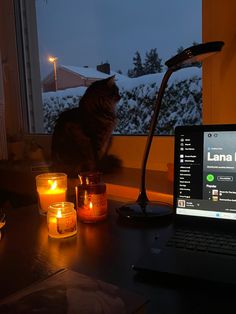 a laptop computer sitting on top of a desk next to candles and a cat looking out the window