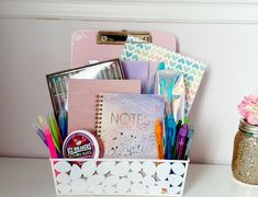 a white container filled with lots of office supplies next to a pink flower on top of a desk