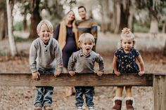 three young children standing on a wooden fence in the woods with their parents behind them