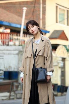 a woman in a trench coat and black dress is standing on the street with her handbag