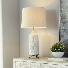 a white lamp sitting on top of a dresser next to a painting and potted plant