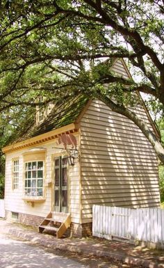 a small yellow house sitting next to a white picket fence and tree lined street in front of it