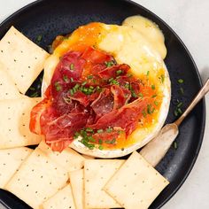 a black plate with crackers, cheese and meat on it next to a wooden spoon