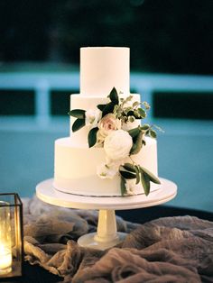 a three tiered white wedding cake with flowers on top and candles in the background