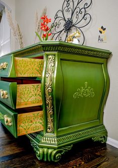 a green painted dresser with ornate designs on the top and bottom, sitting on a hard wood floor in front of a white wall