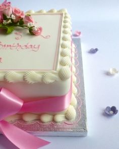 a square birthday cake with pink ribbon and flowers on the top, sitting on a table