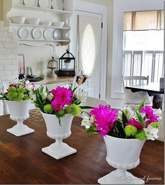 three white vases filled with flowers sitting on top of a wooden dining room table