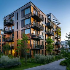 This image showcases a modern Scandinavian apartment building featuring natural wood and dark metal accents. Large windows and glass balconies with black railings offer ample outdoor space. The landscaped surroundings include lush greenery, trees, and a paved pathway, creating a serene environment. This stylish apartment complex combines contemporary design with natural materials for a warm and inviting look. Perfect for those seeking a blend of elegance and modernity in apartment living. Apartment Complex Design, Apartment Building Landscape, Cute Apartment Complex Exterior, Small Apartment Complex Exterior, Wood Apartment Building, Tudor Apartment Building, Small Apartment Complex, Timber Apartment Building