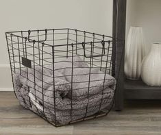 a wire basket with two towels in it on the floor next to a shelf and vase