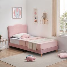 a pink bed in a white room with two stuffed animals on the floor and a window