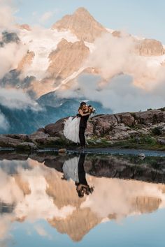 a bride and groom standing in front of a mountain lake with their arms around each other