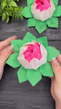 two hands holding origami flowers on top of a wooden table with greenery