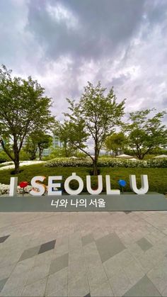 a sign that says seoul on it in front of some trees and bushes with clouds overhead