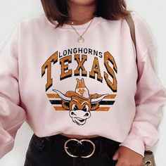 Relive The Classic Ut Austin Days With Our 'Retro Longhorn' Sweatshirt, Showcasing A Vintage Design Of Bevo, The Beloved Longhorns Mascot. Offered In A Wide Range Of Colors And Sizes Exclusively On Our Website, This Piece Is A Nod To The Rich Legacy Of The University. Proudly Designed By Our Small Business, It's A Must-Have For Every True Longhorn Enthusiast. Retro Pink Sweatshirt With Graphic Print, Retro Pink Graphic Print Sweatshirt, Pink Crew Neck Top For Fans, Retro Pink Sweatshirt For Fall, Pink Cotton Sweatshirt For Fan Merchandise, Pink Crew Neck Top For College, Pink Casual Sweatshirt For Fan Merchandise, Pink Letter Print Tops For College, Pink Long Sleeve Sweatshirt For Fan Merchandise