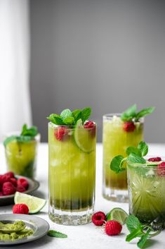 three glasses filled with green drinks and raspberries on top of a white table