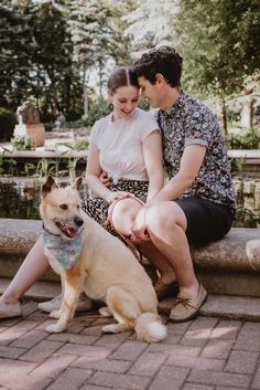 a man and woman sitting next to a dog on a brick walkway with trees in the background