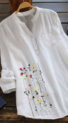 a white shirt with flowers embroidered on it sitting on a wooden bench next to a book