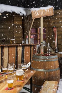 a wooden table topped with lots of glasses filled with beer next to barrels covered in snow