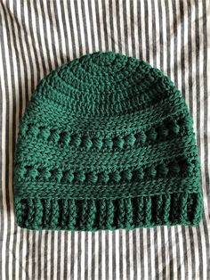 a green crocheted hat laying on top of a striped bed sheet next to a white and black stripped pillow