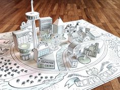 a paper cut out of a city with buildings and trees on the ground, in front of a wooden floor