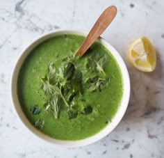 a white bowl filled with green soup next to a slice of lemon and a wooden spoon