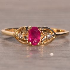 Dos Avenida: Oval Brilliant Cut Ruby And Natural Diamond Heart Ring In Yellow Gold. The Ring Showcases A Striking Oval Brilliant Cut Purplish-Red Natural Ruby Center, Radiating With Strong Saturation. Flanking The Center Are Two Round Cut Natural Diamonds, Each Delicately Set At The Cleft Of A Heart-Shaped Design That Adorns The Top Of The Tapered Shank. Crafted In Yellow Gold, This Ring Is Currently A Finger Size 6 And Can Be Adjusted To Any Finger Size For An Additional Charge Upon Request. Elegant Heart Shaped Ruby Ring In Yellow Gold, Gold Heart Ring With Oval Gemstone, Elegant Yellow Gold Heart Ring With Gemstone, Gold Heart Ring With Gemstone, Gold Birthstone Ring For Valentine's Day Formal, Yellow Gold Heart-shaped Formal Birthstone Ring, Yellow Gold Heart-shaped Birthstone Ring For Formal Occasions, Anniversary Yellow Gold Ruby Ring With Gemstone Accents, Heart-shaped Yellow Gold Birthstone Ring For Formal Occasions