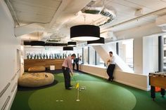 two men playing golf in an office with green carpet and white walls, while another man is on the other side of the room