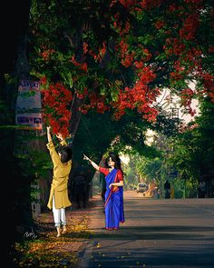 two women standing on the side of a road near trees with red flowers in their hands