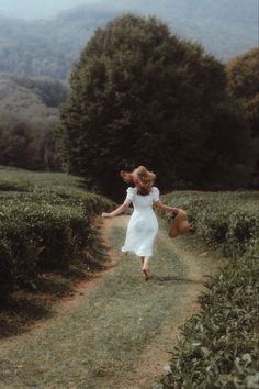 a woman in a white dress is running down a dirt road with trees and bushes behind her