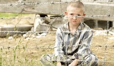 a young boy sitting on the ground in front of a barbed wire fence and looking at the camera