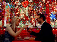 a man and woman sitting at a table with wine glasses in front of christmas decorations