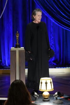 an older woman standing at a podium in front of a blue curtain with a lamp on it