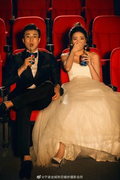 a man and woman sitting on red chairs in front of an empty theater with drinks